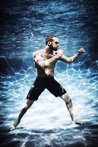 Underwater shot of a muscular adult male striking a fighting pose with bare torso, black shorts, and neutral expression. 