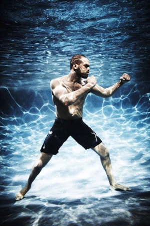 Underwater shot of a muscular adult male striking a fighting pose with bare torso, black shorts, and neutral expression. 
