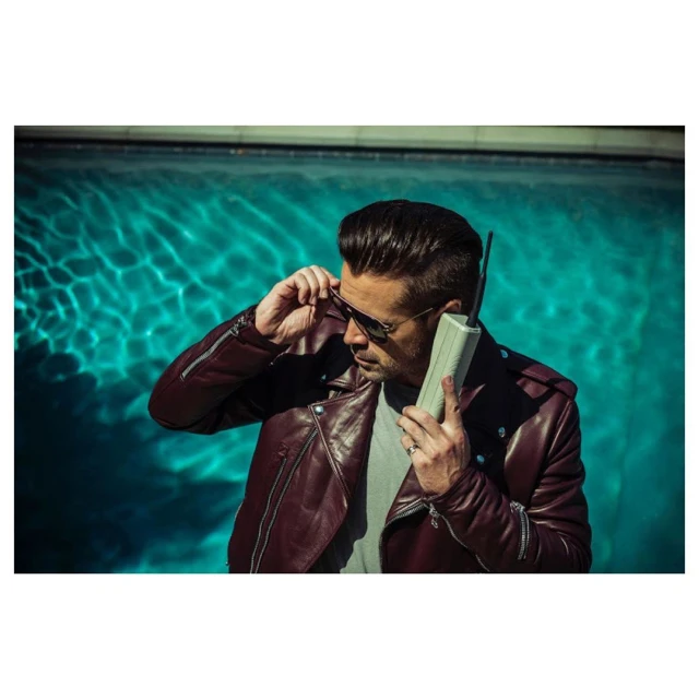 Adult male in sunglasses and a burgundy leather jacket poses by a swimming pool while holding a retro cellular phone to his ear.
