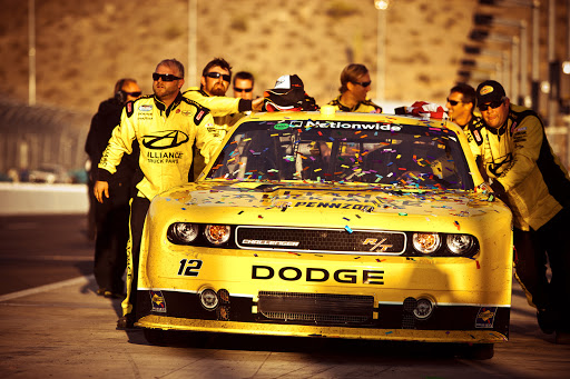 A yellow Dodge race car surrounded by a pit crew celebrating a win on the racetrack with confetti. The desert hills are in the background.