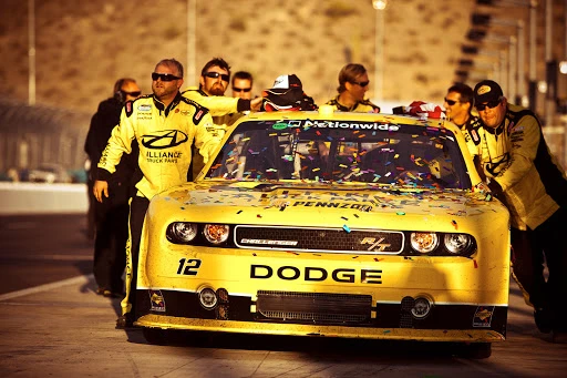 A yellow Dodge race car surrounded by a pit crew celebrating a win on the racetrack with confetti. The desert hills are in the background.