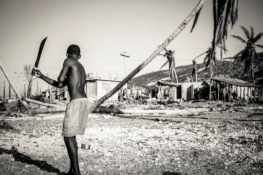 Black-and-white photograph of a young person holding a machete outdoors amid damaged palm trees and buildings.