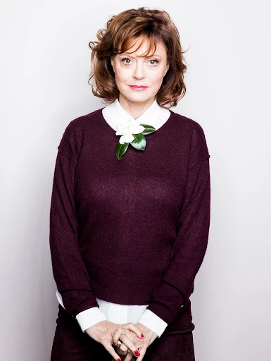 Portrait of Susan Sarandon wearing a burgundy sweater over a white shirt with a white flower pinned to the collar against a white background.