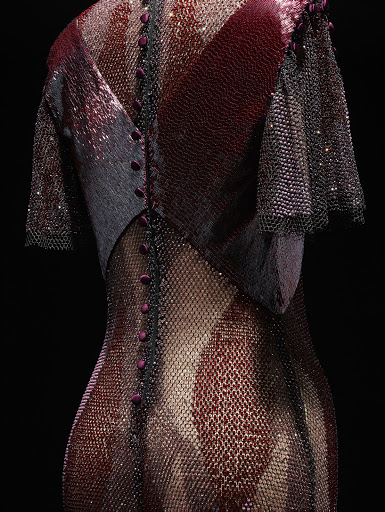 Back view of a shimmering, intricately designed formal dress with a gradient effect, adorned with buttons and delicate sleeves on black backdrop.