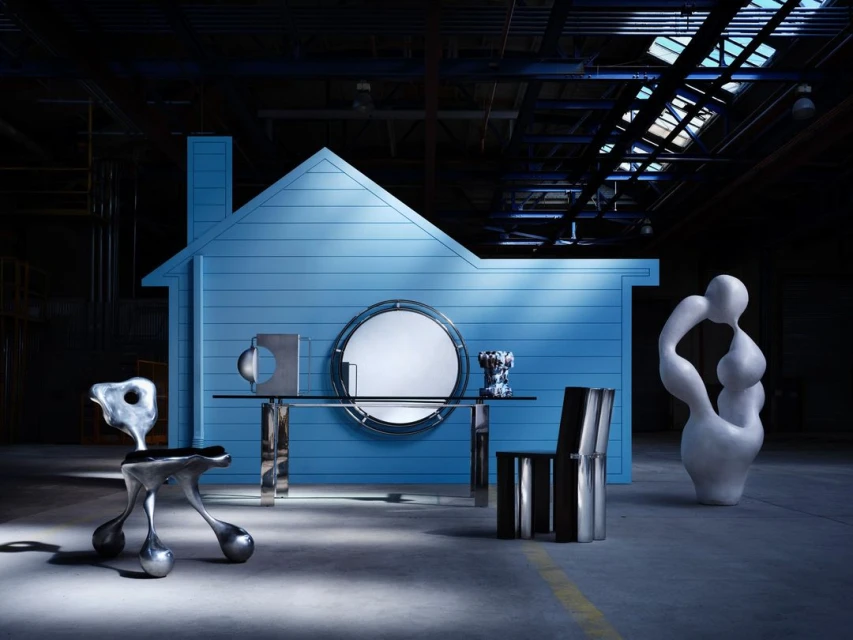 Modern furniture arranged in front of a blue house structure in a studio, featuring chairs, a table, and a sculpture.