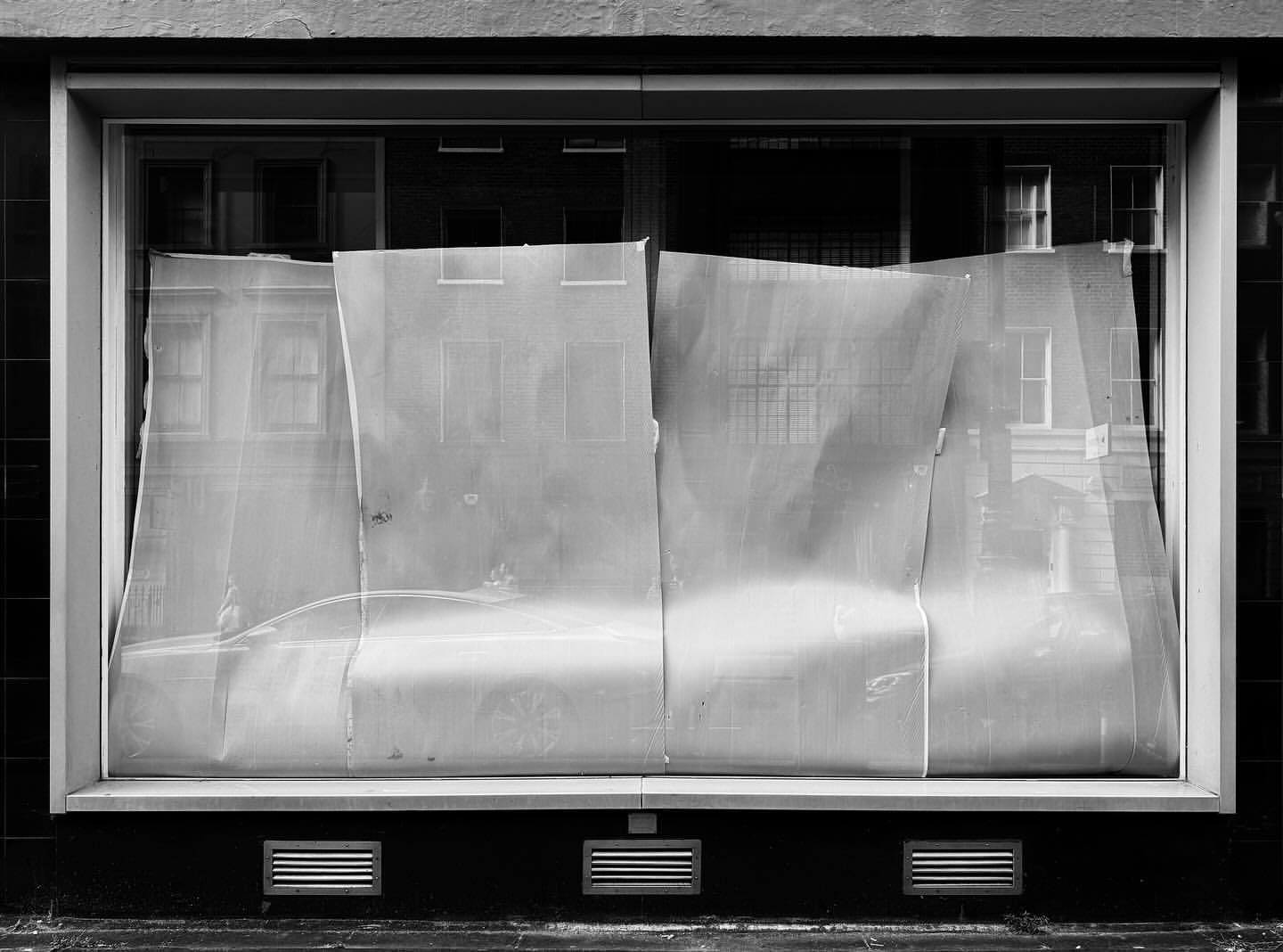 Black and white architectural photograph of a storefront window covered with sheer fabric, reflecting the surrounding urban buildings.