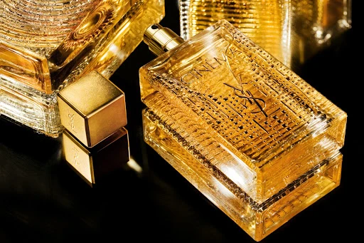Golden rectangular perfume bottle with a textured glass pattern and metallic cap resting on a dark reflective surface