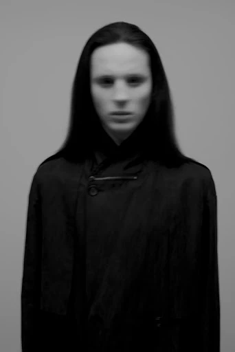 Black and white studio portrait of a person with long dark hair wearing a dark overcoat against a neutral grey background.