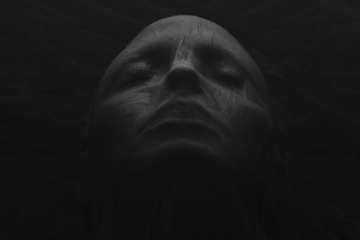 Monochromatic portrait of an adult with eyes closed, face covered in textured fabric, set against a dark, shadowy background.