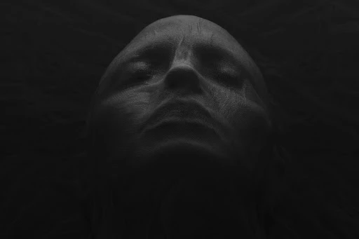 Monochromatic portrait of an adult with eyes closed, face covered in textured fabric, set against a dark, shadowy background.