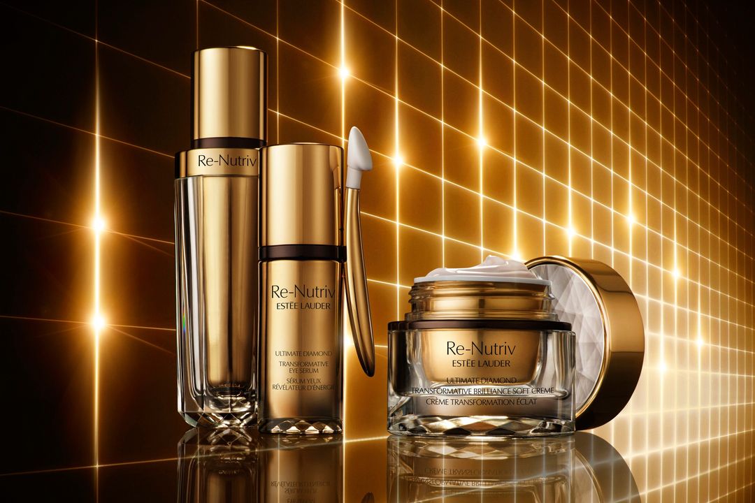 Three gold and glass Re-Nutriv Estee Lauder skincare product containers sit on a reflective surface against a background of lit grid lines.