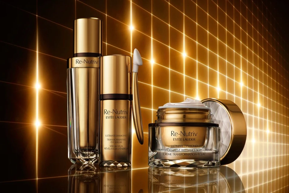 Three gold and glass Re-Nutriv Estee Lauder skincare product containers sit on a reflective surface against a background of lit grid lines.