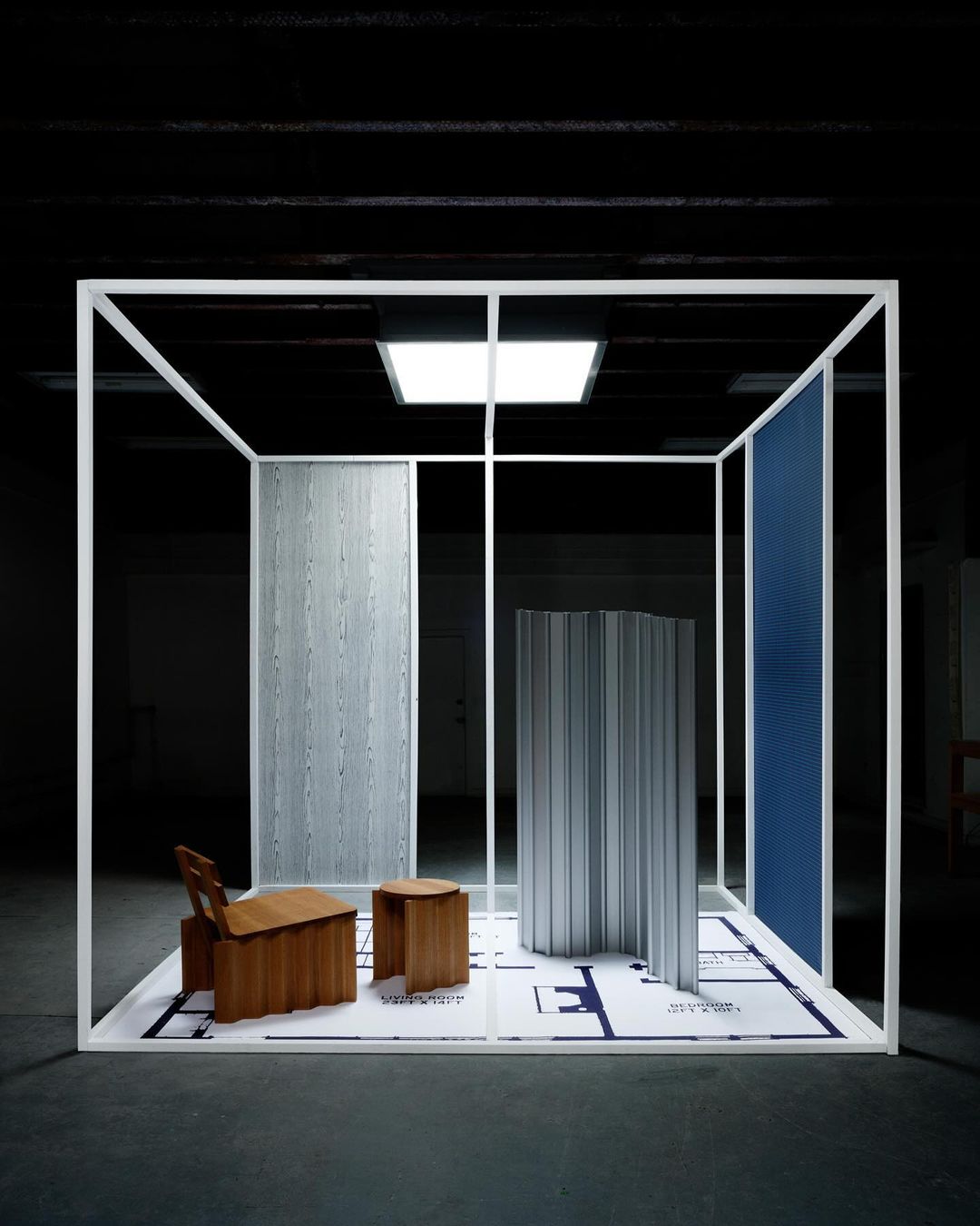 Interior design installation featuring deconstructed furniture pieces, partitions, and a blueprint floor within a white frame structure, indoors.