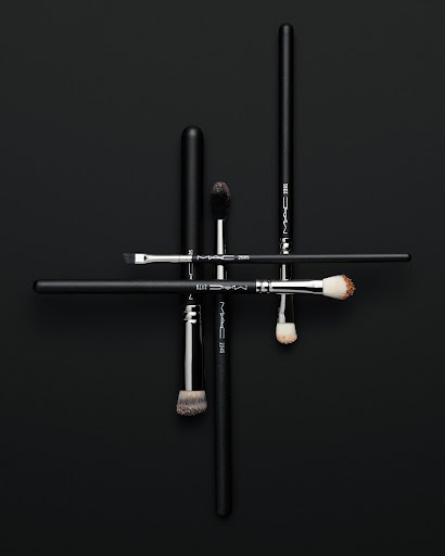 Overhead studio shot of a group of black handled makeup brushes arranged in an abstract linear pattern on a black background.