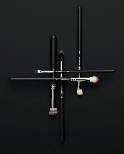 Overhead studio shot of a group of black handled makeup brushes arranged in an abstract linear pattern on a black background.