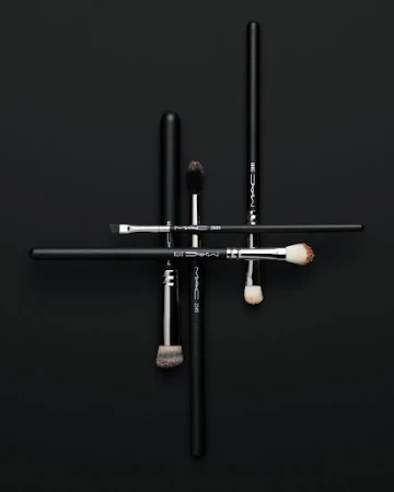 Overhead studio shot of a group of black handled makeup brushes arranged in an abstract linear pattern on a black background.