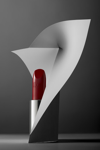 Close-up studio shot of red lipstick in a silver tube, nestled in a paper sculpture, set against a gradient dark grey backdrop.