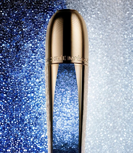 Gold Orchidee Imperiale skincare product stands against a background of blue and white spheres, highlighting luxury and beauty.