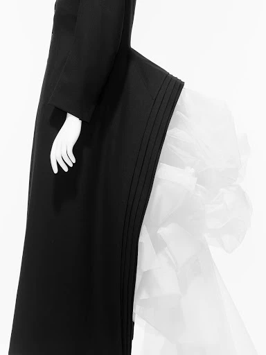 Black and white studio shot of a mannequin wearing a black dress with a white ruffled accent, minimal background, and neutral lighting.