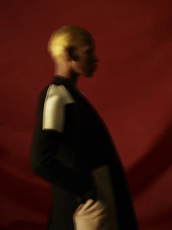Motion blurred profile portrait of a black man with dyed blonde hair wearing a dark jacket against a red backdrop