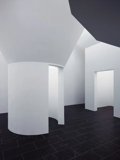 Minimalist interior featuring white walls, a curved architectural element, black tiled floor, and natural light entering the room.