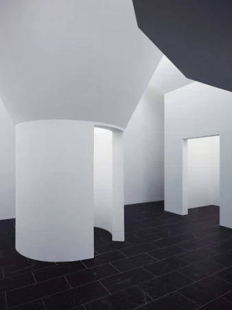 Minimalist interior featuring white walls, a curved architectural element, black tiled floor, and natural light entering the room.