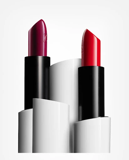 Close-up studio shot of two lipsticks, one red and one dark berry, standing against an arrangement of white cylinders.