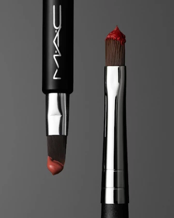 Two MAC makeup brushes with cream lipstick on bristles against grey background