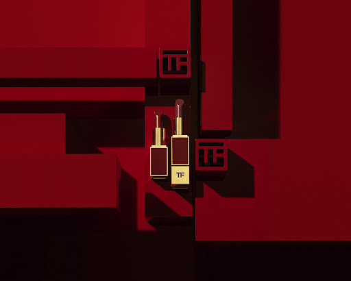 Two red and gold Tom Ford lipsticks sit atop red geometric structures, creating a visually striking beauty product still life.