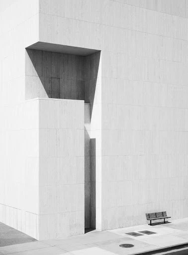 Minimalist black and white architectural photograph features stark geometric building exterior with shadows and a bench.