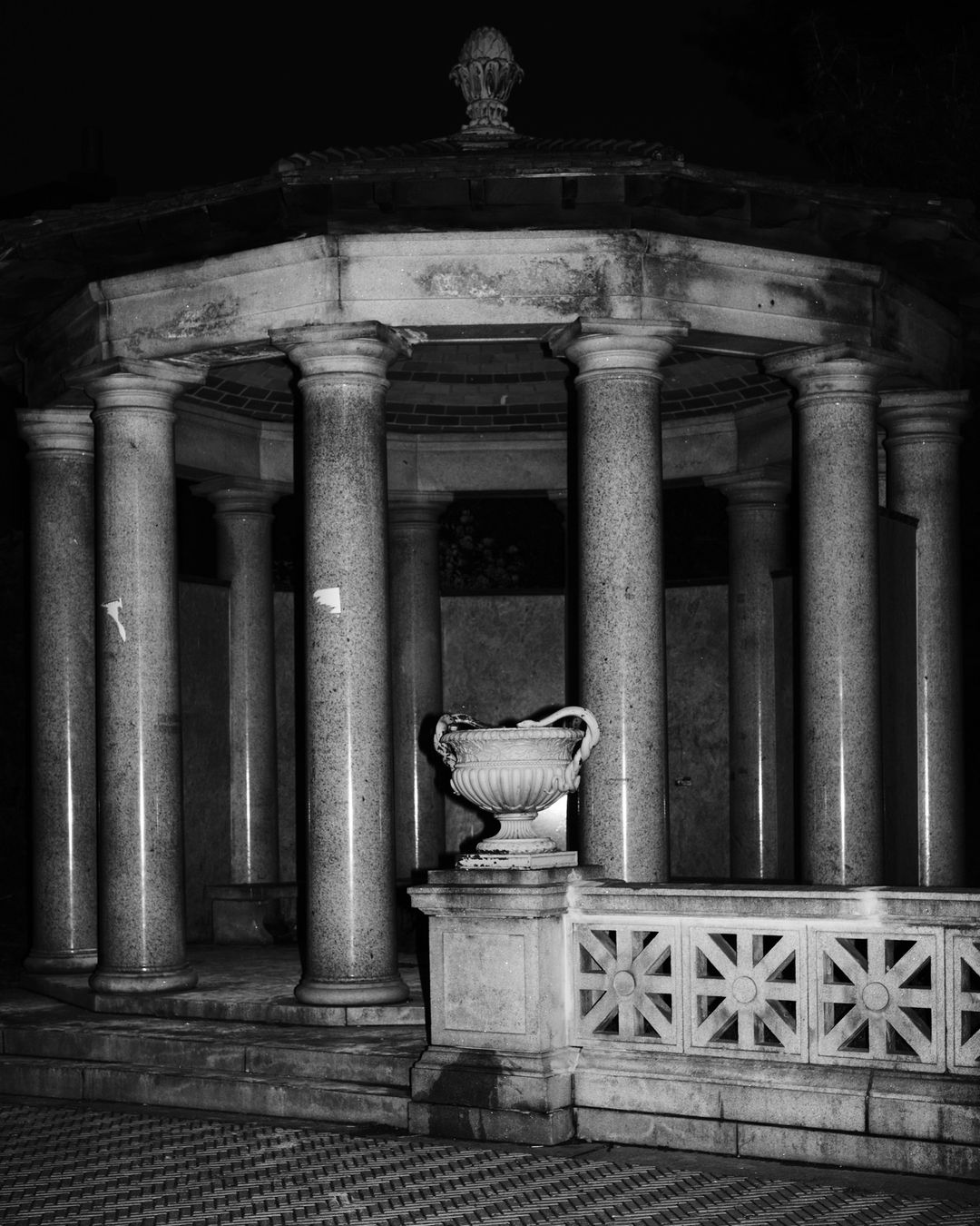 Black and white image of a marble gazebo with columns and an ornate vase on a platform with a geometric tile floor.