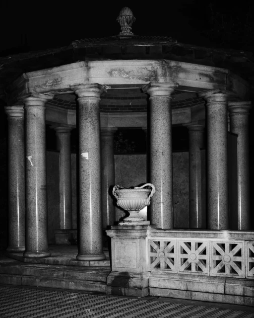 Black and white image of a marble gazebo with columns and an ornate vase on a platform with a geometric tile floor.