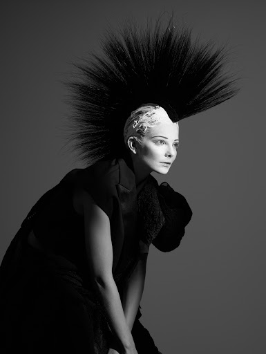 Monochrome portrait of a woman with an avant-garde black and white hairstyle and dark clothing against a grey studio backdrop.