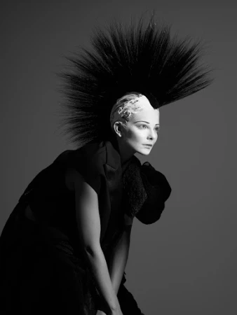 Monochrome portrait of a woman with an avant-garde black and white hairstyle and dark clothing against a grey studio backdrop.