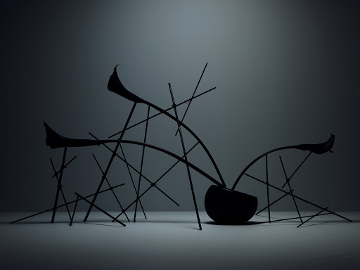 Dark botanical arrangement in a round vessel supported by slender geometric sticks against a plain grey gradient background.