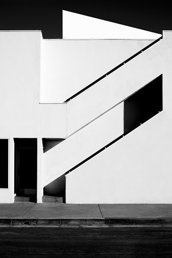 Black and white architectural photograph of a stark white building with angled staircases and strong shadows on a sunny day.