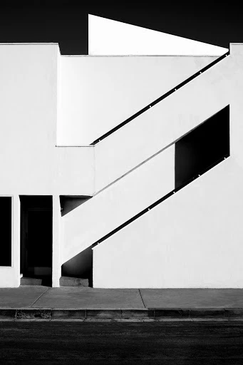Black and white architectural photograph of a stark white building with angled staircases and strong shadows on a sunny day.