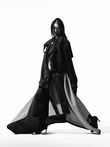 A solo female fashion model in a flowing dress and trench coat poses in a studio with bright lighting. The image is black and white.