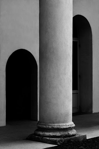 Monochrome photo of a classical column, arched doorway, and a building exterior with a dark shadowed passage, creating an architectural scene.
