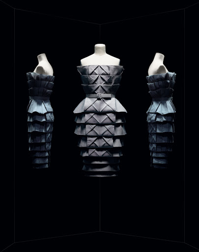 Three mannequins in a staggered arrangement, each wearing a gray, layered origami-style dress against a stark black backdrop.