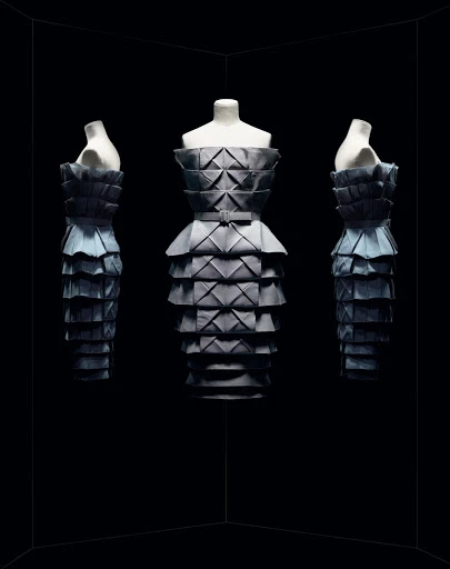 Three mannequins in a staggered arrangement, each wearing a gray, layered origami-style dress against a stark black backdrop.
