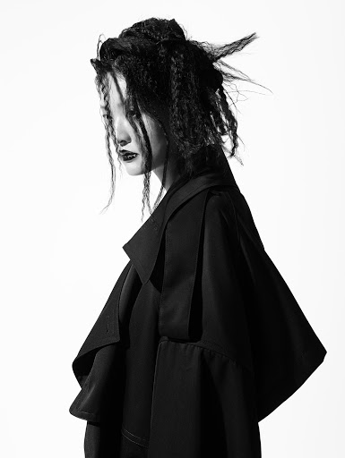 Black-and-white portrait of a slender young woman with intricate black hair, dark lipstick, and a black coat against a white backdrop.