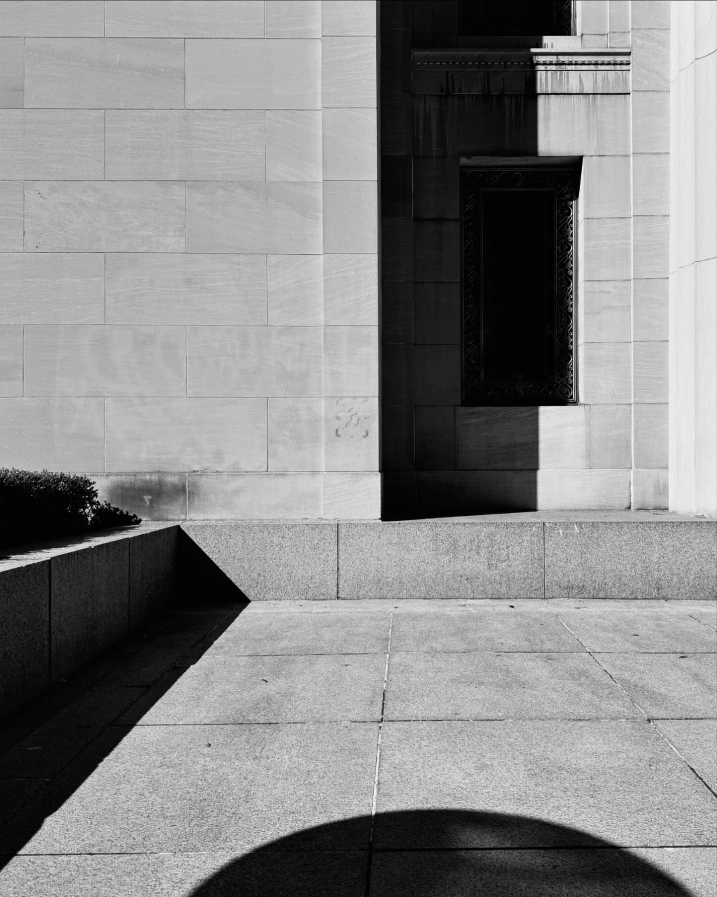 Monochrome image of a stone building facade with a dark window, concrete steps, and shadows creating a geometric composition.