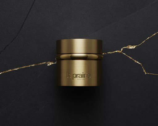 A close-up studio shot features a gold-colored La Prairie beauty product against a dark background with a gold vein accent.