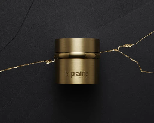 A close-up studio shot features a gold-colored La Prairie beauty product against a dark background with a gold vein accent.
