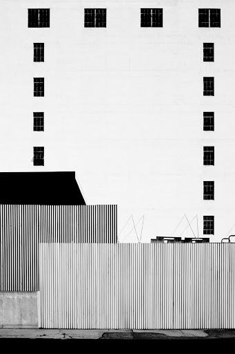 Minimalist black-and-white architectural photograph of a tall building with windows behind corrugated metal fencing along the sidewalk.
