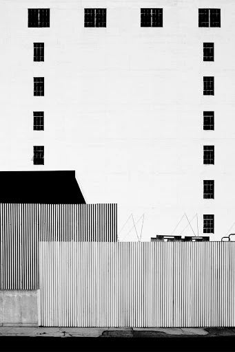 Minimalist black-and-white architectural photograph of a tall building with windows behind corrugated metal fencing along the sidewalk.