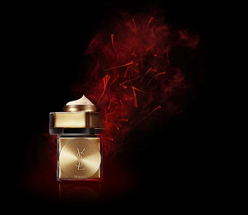 Elevated close-up of stacked gold YSL beauty product containers against a dark background with a burst of red powder.