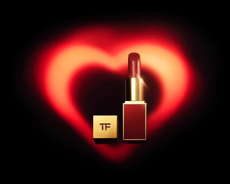 Tom Ford lipstick in a red and gold tube, displayed against a dark background with a soft, glowing red heart shape behind it.