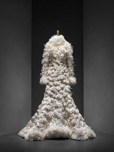 Full-length view of a couture ivory coat with textured rose appliqués, presented on a mannequin against a dark, graduated background.